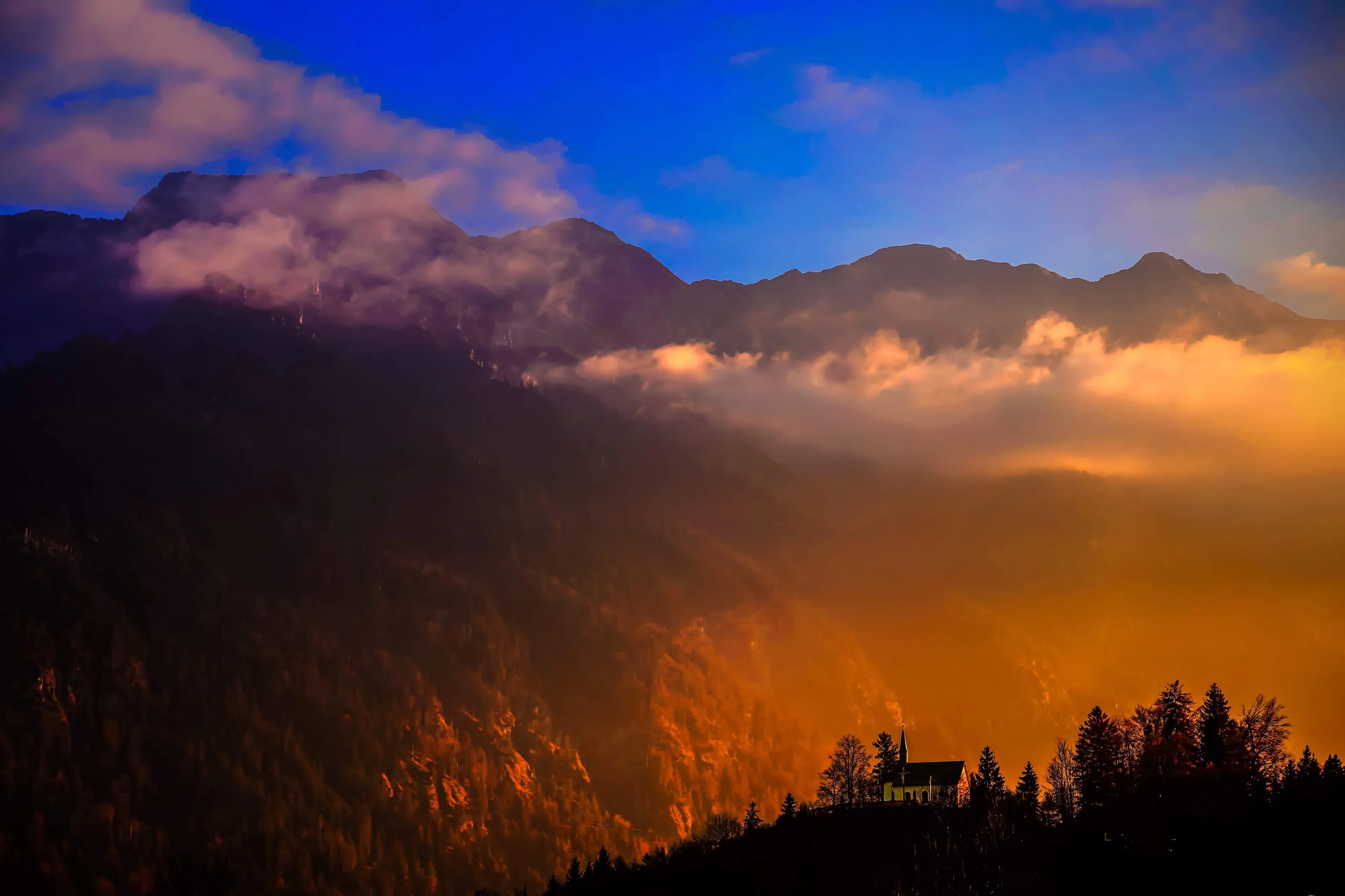 The beginning of golden hour a little white church sit at atop of a hill, with the Bavarian alps as a backdrop.
The image goes from golden blue skies to deep black.