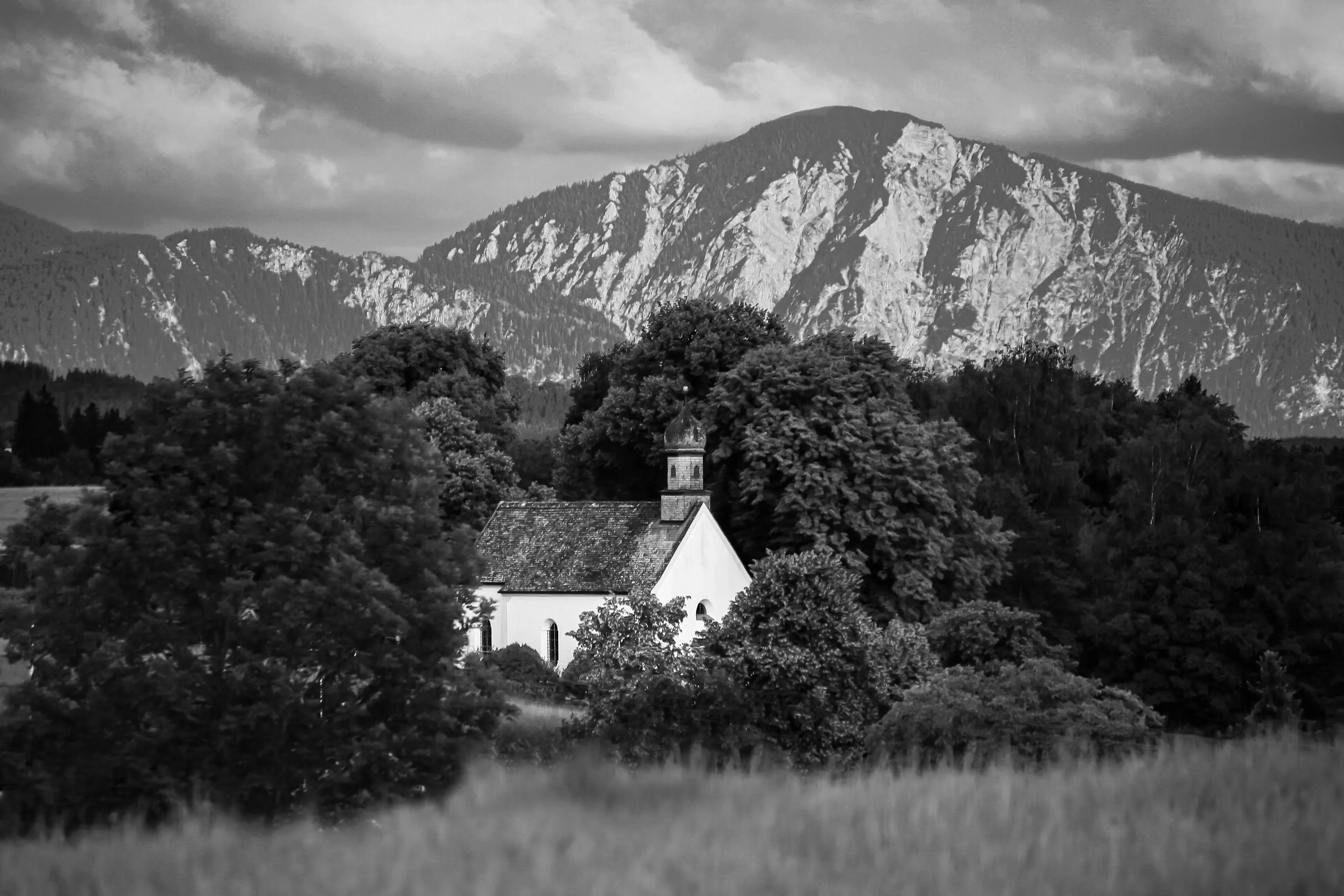 A dramatic black and white landscape featuring a small, white chapel with a steeple and onion dome.
Background is a massive rugged mountain with light colored rocky faces rises beneath a cloudy, moody sky. The foreground shows a grassy field, adding depth to the scene.